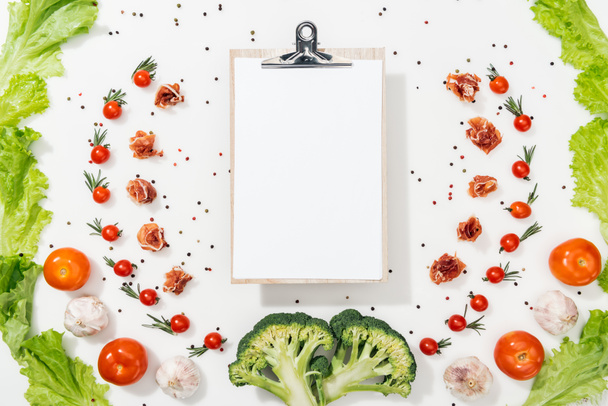 top view of empty clipboard among tomatoes, lettuce leaves, prosciutto, broccoli, spices and garlic - Fotó, kép