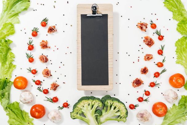 top view of black clipboard among tomatoes, lettuce leaves, prosciutto, broccoli, spices and garlic - Fotó, kép