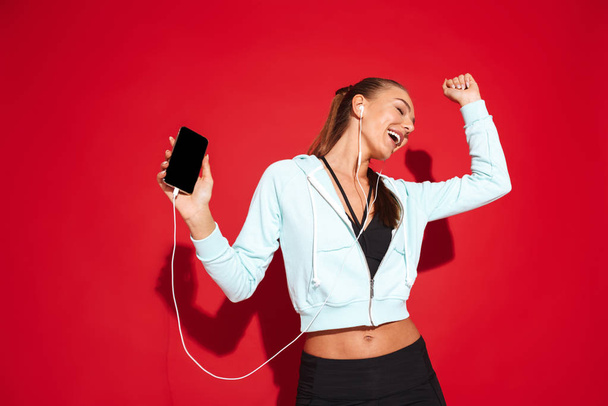 Portrait of a beautiful fit young sportswoman standing over red background, listening to music with earphones, holding mobile phone - Fotoğraf, Görsel