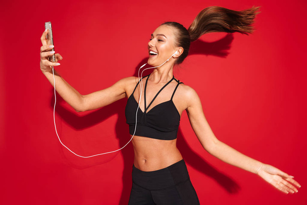 Portrait of a beautiful fit young sportswoman jumping over red background, listening to music with earphones, holding mobile phone - Φωτογραφία, εικόνα