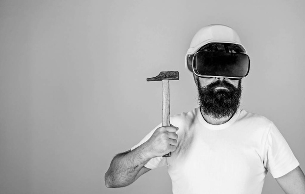 Hipster on serious face wear helmet, hold hammer in virtual reality. Man with beard in VR glasses hold hammer, light blue background. Virtual renovation concept. Guy builder with head mounted display - Foto, Imagen