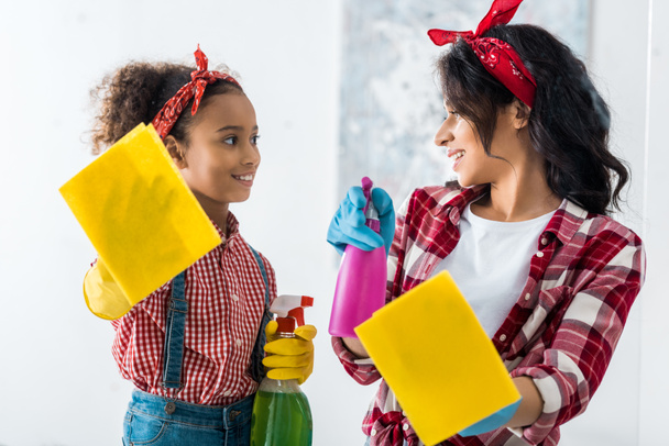 sorridente afro-americano mãe e filha com trapos amarelos e spray garrafas casa de limpeza
 - Foto, Imagem