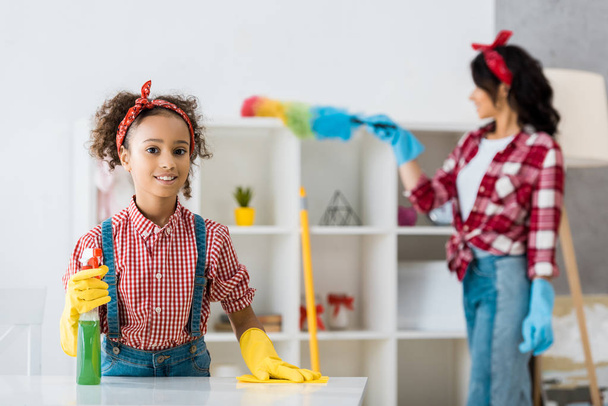 selective focus of smiling african american child cleaning table while mother dusting shelving unit - Фото, изображение