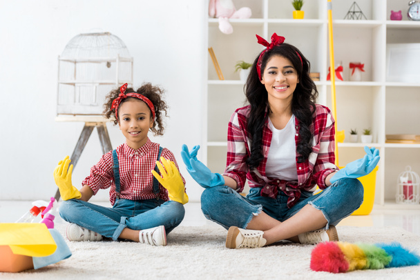 smiling african american mom and daughter and plaid shirts and bright rubber gloves sitting in lotus pose  - Φωτογραφία, εικόνα