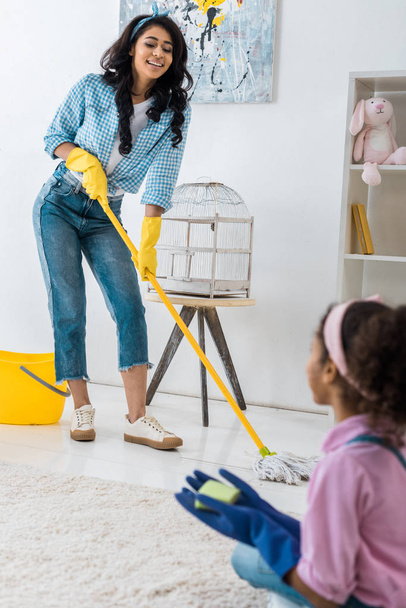smiling african american woman washing floor with mop while daughter sitting on carpet - Φωτογραφία, εικόνα