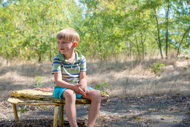 The boy sits on an old bench, in an abandoned park and looks around with a sad face. - Photo, Image