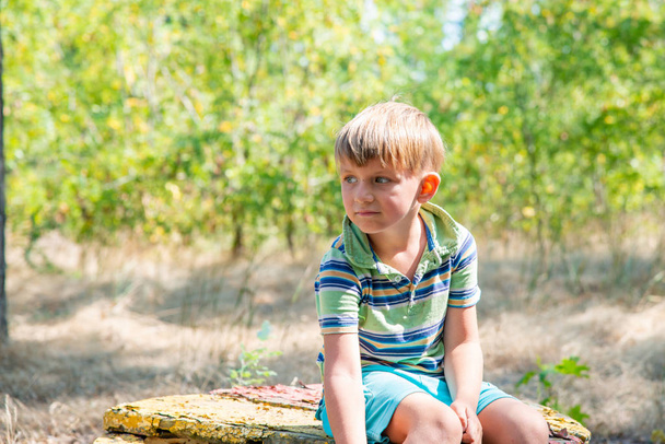 Le garçon est assis sur un vieux banc, dans un parc abandonné et regarde autour de lui avec un visage triste
. - Photo, image