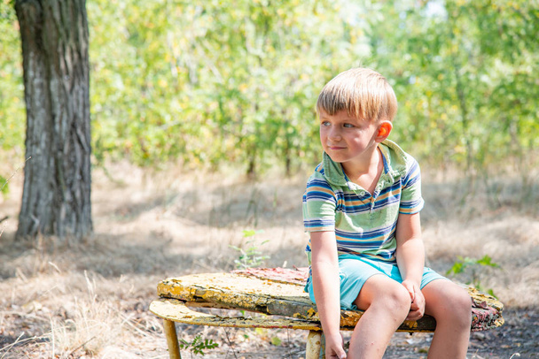 O menino se senta em um banco velho, em um parque abandonado e olha em volta com um rosto triste
. - Foto, Imagem