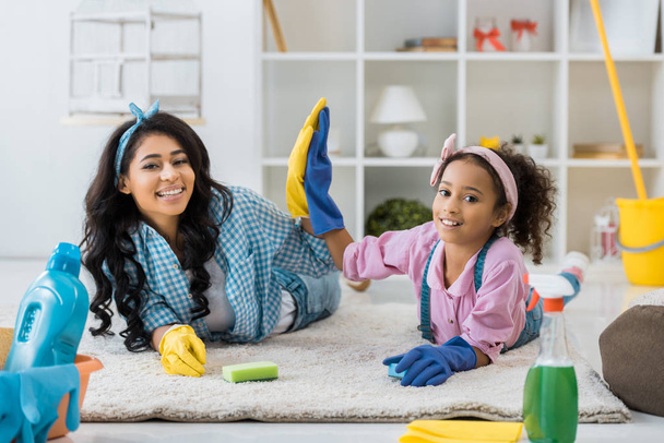 smling african american mother and child in bright rubber gloves giving high five while lying on carpet - Photo, Image