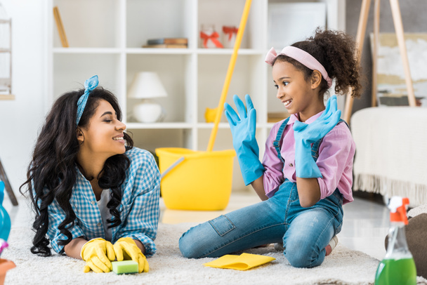smiling african american mom and daughter in bright rubber gloves talking while resting on carpet - Foto, afbeelding