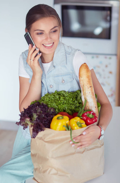 Smiling woman with mobile phone holding shopping bag in kitchen - Фото, зображення