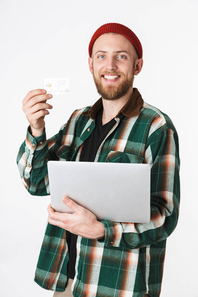 Portrait of unshaved man holding silver laptop and credit card while standing isolated over white background - Fotografie, Obrázek