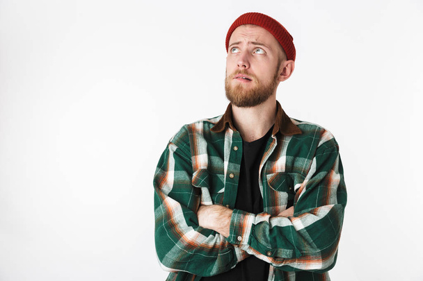 Portrait of young bearded guy wearing hat and plaid shirt looking up while standing isolated over white background - Valokuva, kuva