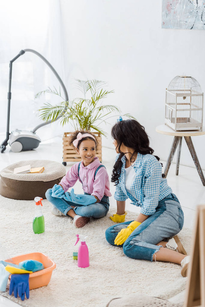 african american american mother and daughter in bright rubber gloves talking while sitting on carpet - Fotó, kép