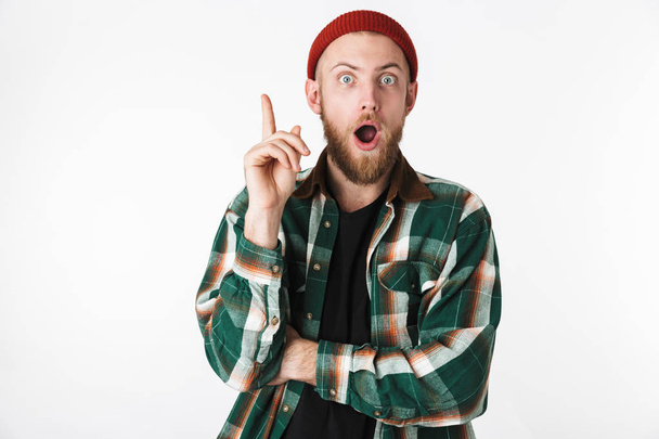 Portrait of surprised bearded guy wearing hat and plaid shirt pointing finger upward while standing isolated over white background - Foto, immagini