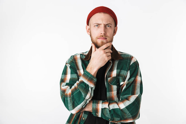 Portrait of european guy wearing hat and plaid shirt touching his chin while standing isolated over white background - Foto, afbeelding