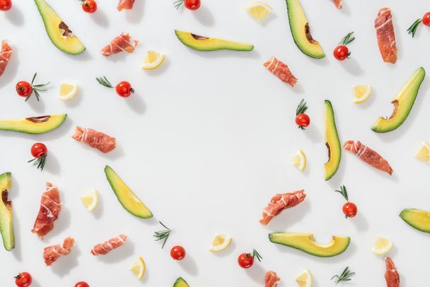 flache Lage von Prosciutto in der Nähe von in Scheiben geschnittenen Avocados, Zitronen und Kirschtomaten auf weißem Hintergrund - Foto, Bild