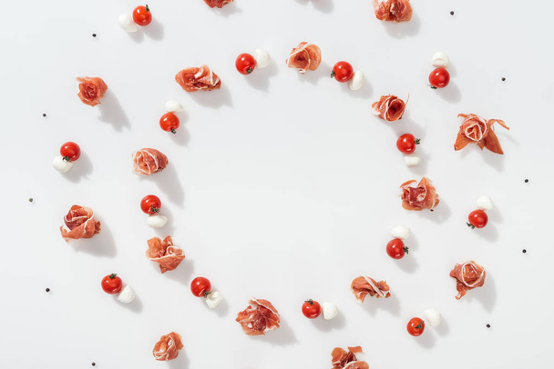 flat lay of tasty prosciutto near red cherry tomatoes, mozzarella cheese and peppercorns on white background - Photo, Image