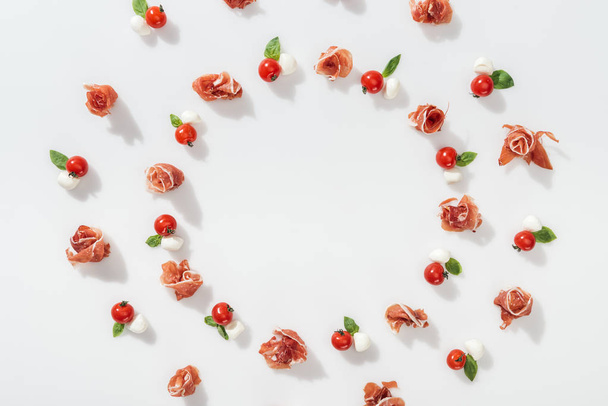 plat leggen van smakelijke prosciutto in de buurt van rode cherry tomaten, mozzarella kaas en groene Basilicum bladeren op witte achtergrond - Foto, afbeelding