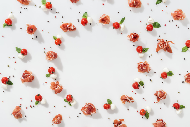 flat lay of prosciutto near red cherry tomatoes, mozzarella cheese and green basil leaves on white background - Photo, Image
