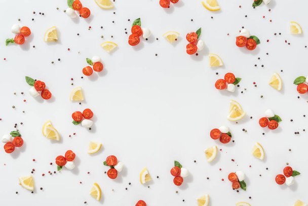 flat lay of red cherry tomatoes, mozzarella cheese, green basil leaves and slices lemons near peppercorns on white background - Photo, Image