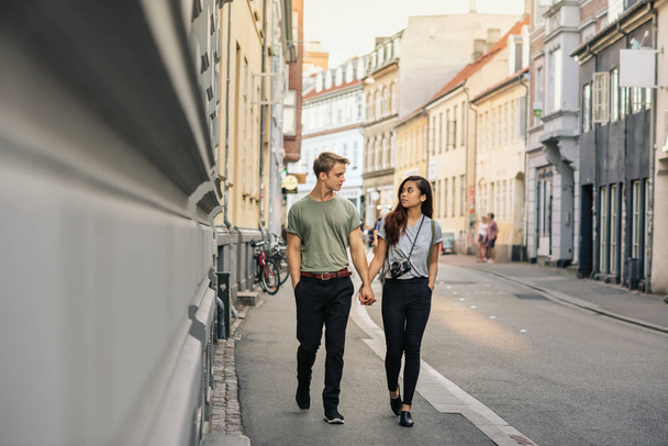 Young couple holding hands and talking together while walking along a sidewalk in the city - Zdjęcie, obraz