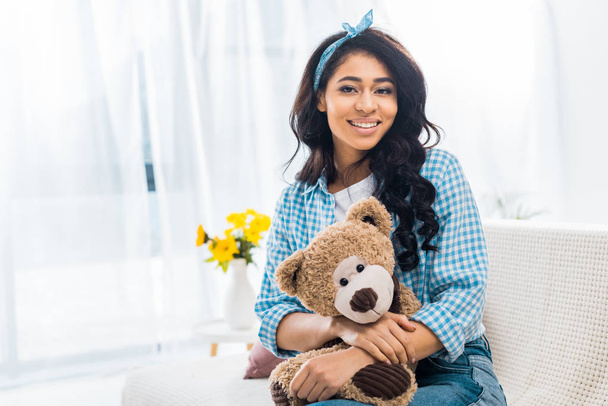 beautiful african american woman sitting on sofa and holding teddy bear - Foto, Imagen