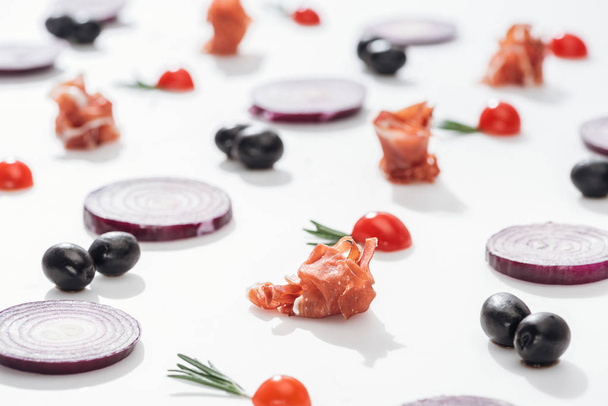 selective focus of raw prosciutto near cherry tomatoes with rosemary twigs near red onion rings and black olives on white background - 写真・画像