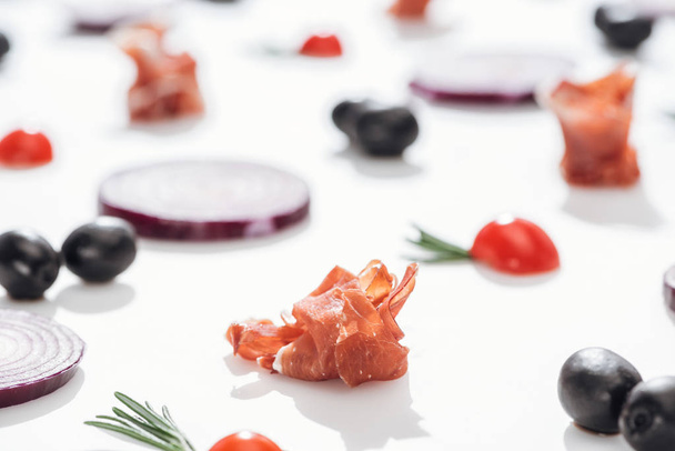 selective focus of delicious prosciutto near cherry tomatoes with rosemary twigs and red onion rings on white background - Fotografie, Obrázek