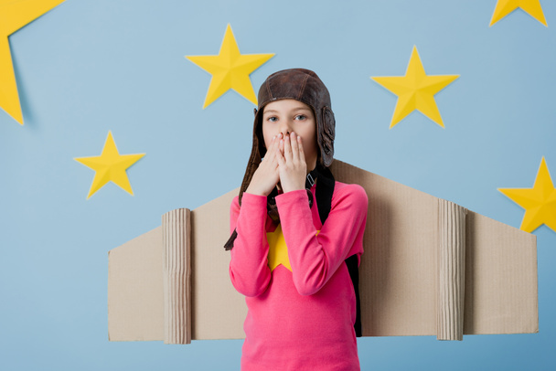 Surprised child with cardboard wings covering mouth with hands on blue starry background - Foto, afbeelding