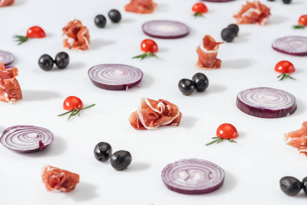 selective focus of red onions near prosciutto and cherry tomatoes with rosemary twigs on white background - Fotó, kép