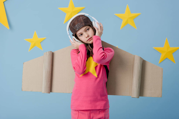 Niño soñador con alas de cartón escuchando música sobre fondo azul estrellado
 - Foto, Imagen