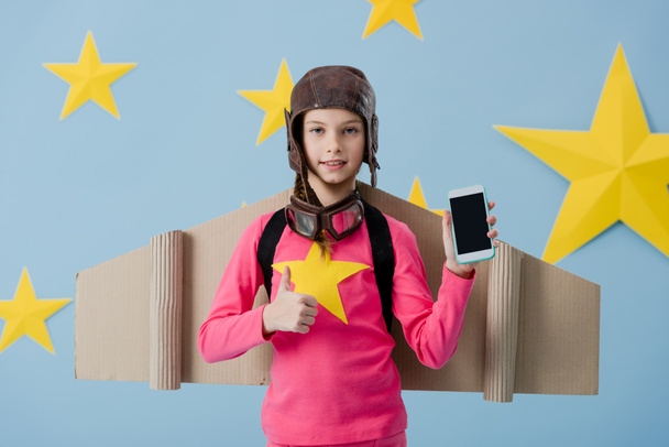 Alegre niño en casco de vuelo sosteniendo teléfono inteligente con pantalla en blanco y mostrando el pulgar hacia arriba sobre fondo estrellado azul
 - Foto, Imagen