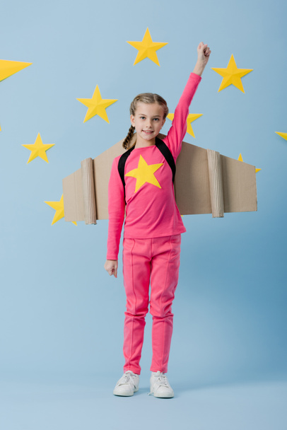 Criança alegre com asas de papelão de pé com punho no fundo azul com estrelas
 - Foto, Imagem