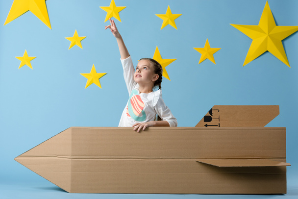 Kid with cardboard rocket pointing with finger at star on blue starry background - Foto, immagini