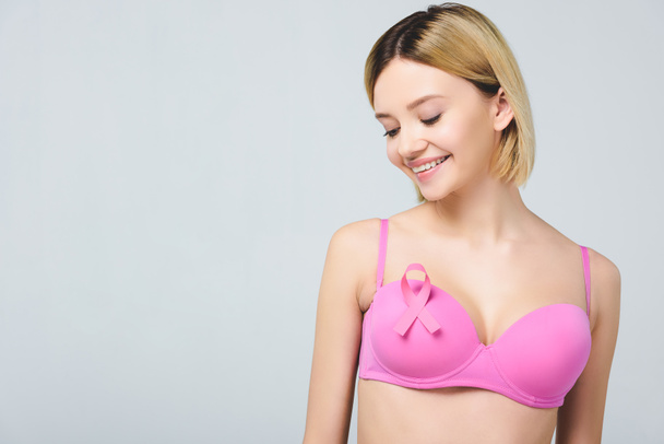 attractive smiling young woman in pink bra with breast cancer awareness ribbon, isolated on grey - Photo, Image