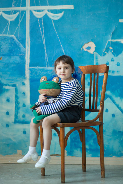 Ein kleines ernstes Mädchen in Rock und gestreiftem T-Shirt sitzt auf einem Stuhl und hält ein altes Spielzeug auf blauem Hintergrund - Foto, Bild