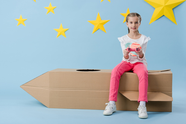 Kid with gamepad sitting on cardboard rocket on blue background with stars - Фото, изображение