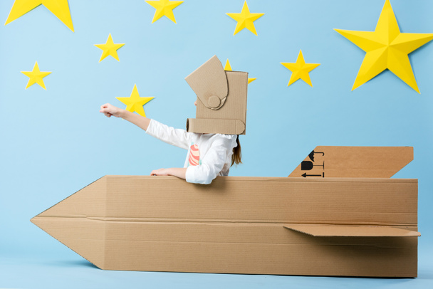 Kid in helmet sitting in cardboard rocket and holding fist up on blue starry background - Photo, Image