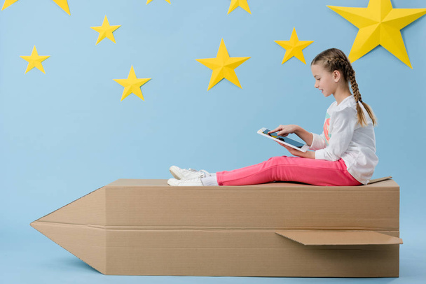 Kind mit Zöpfen sitzt auf Papprakete und nutzt digitales Tablet auf blauem Sternenhintergrund - Foto, Bild