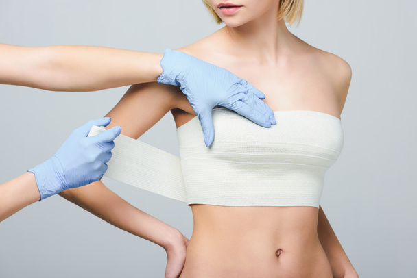 visão recortada de cirurgião plástico envolvendo mama feminina com bandagem, isolado em cinza
 - Foto, Imagem