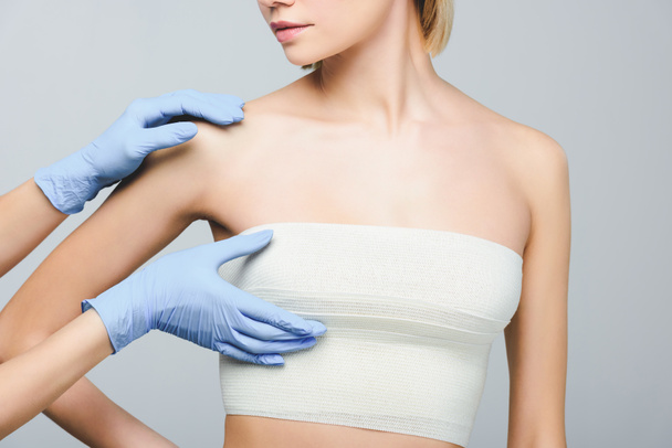 cropped view of plastic surgeon in latex gloves checking female breast in bandage, isolated on grey - Photo, Image