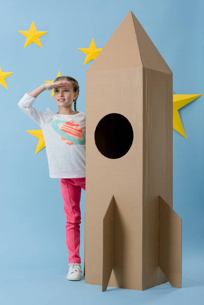 Cheerful kid standing near cardboard rocket and looking away on blue starry background - Foto, afbeelding