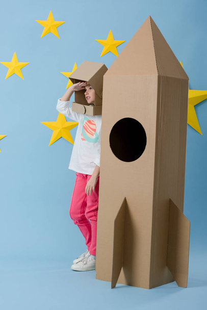 Kind mit Papphelm steht neben Rakete und schaut auf blauem Sternenhintergrund weg - Foto, Bild