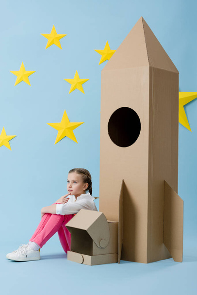 Dreamy kid with helmet sitting near cardboard rocket on blue starry background - Foto, Imagem