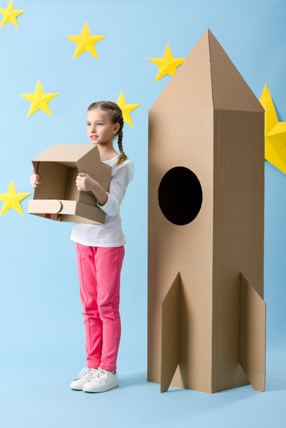 Inspired kid standing near big cardboard rocket and holding helmet on blue starry background - Foto, Imagem