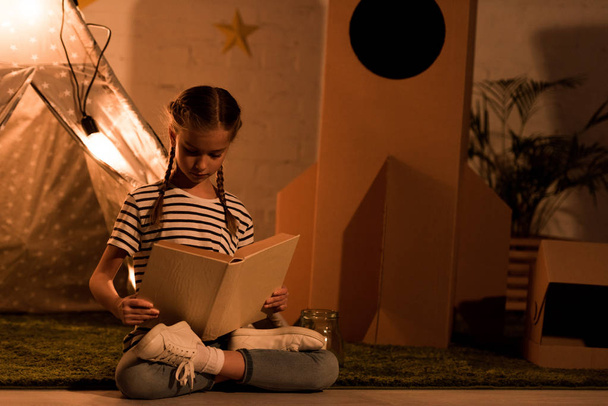 Preteen kind zitten in lotus houding en leesboek in donkere kamer - Foto, afbeelding