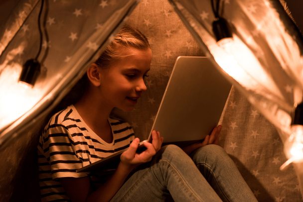 Χαρούμενο παιδί χρησιμοποιώντας το φορητό υπολογιστή σε wigwam στο σπίτι - Φωτογραφία, εικόνα