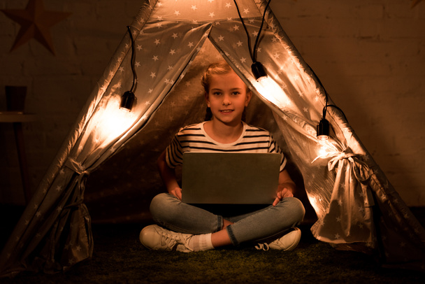Happy kid using laptop while sitting in wigwam in dark room - Photo, image