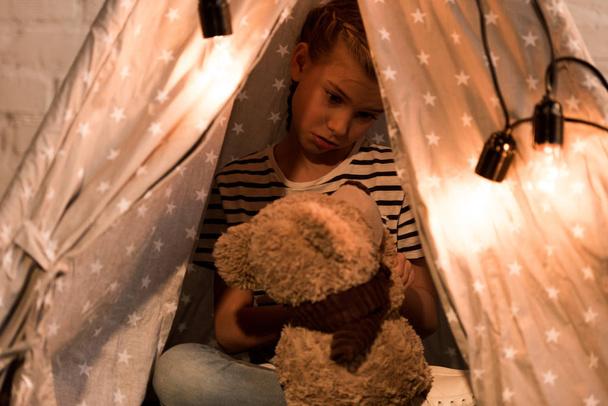 Disappointed kid looking at teddy bear while sitting in wigwam at home - Photo, Image